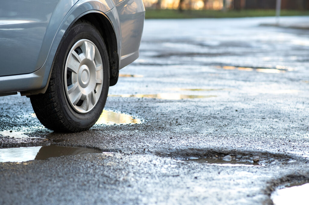 Gros plan sur une roue de voiture sur une route, parsemée de gros nids-de-poule remplis de flaques d'eau de pluie.
