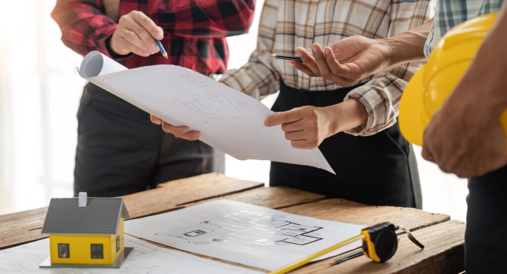 Trois personnes examinent des plans de construction autour d’une table, avec une maquette de maison et un casque de chantier.
